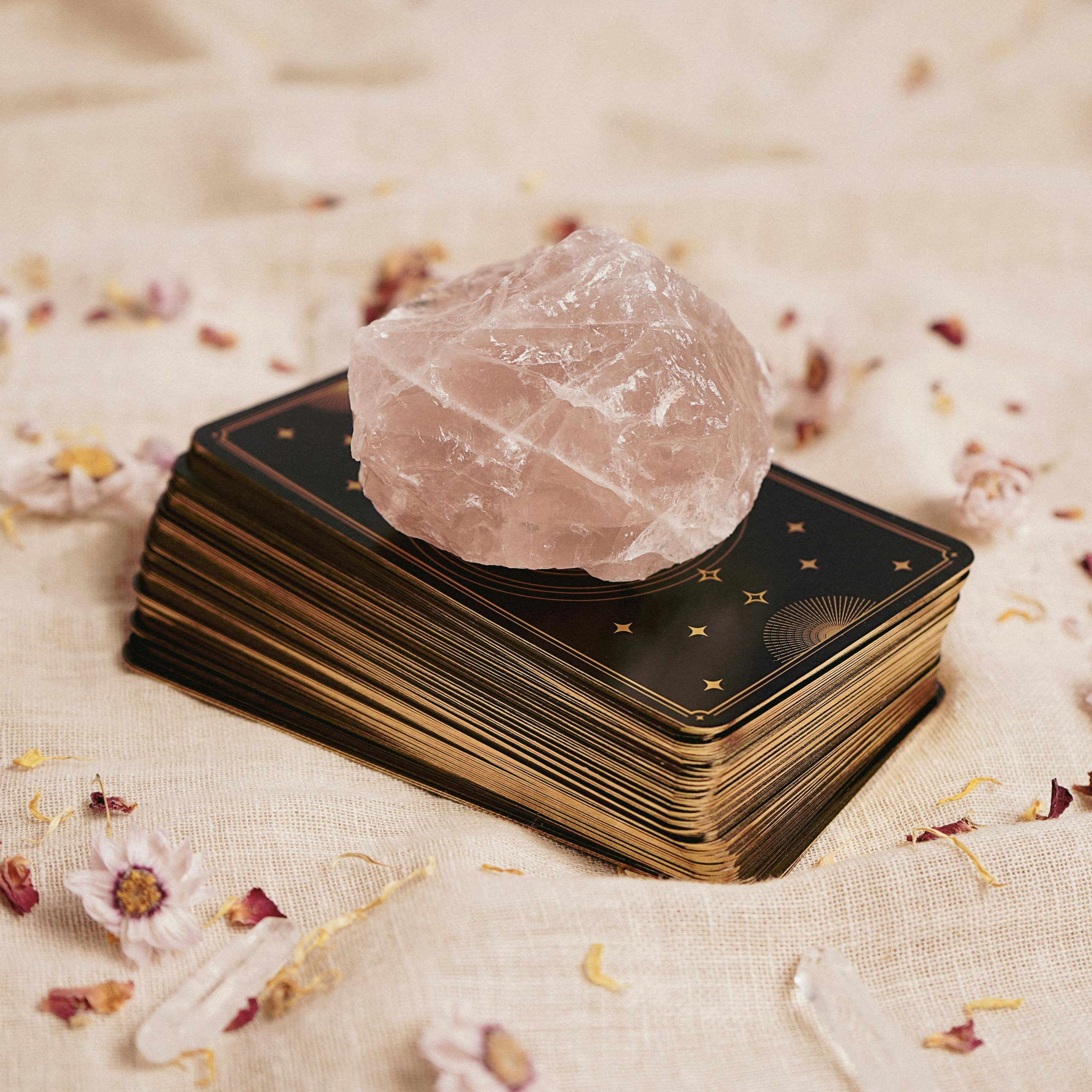 A stack of tarot cards with a rose quartz crystal on top, surrounded by petals on a fabric surface.